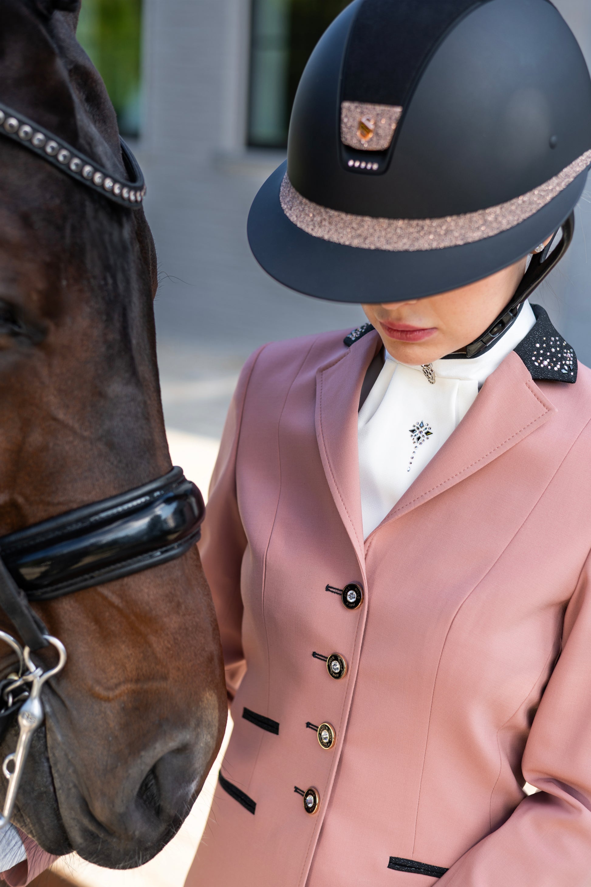 Close-up of model looking down with the head of her horse next to her wearing a competition jacket from JuulC in Blush Rose