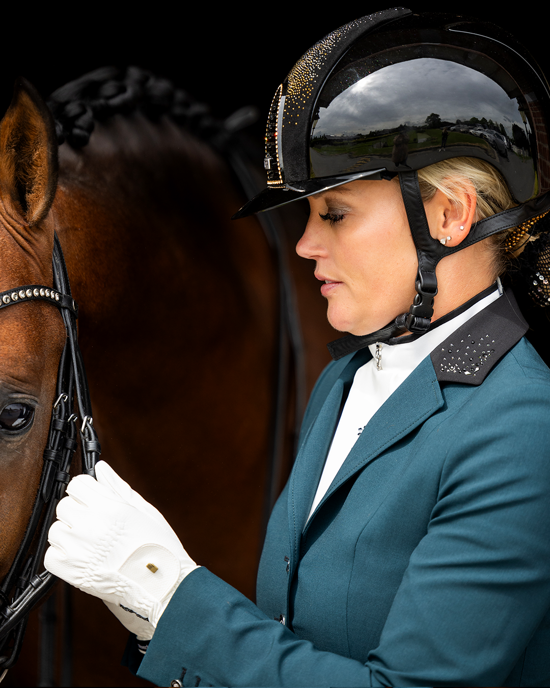 Close-up of model adjusting the bridle of her horse while wearing JuulC' Emerald Women's Show Coat made from sport-tech merino wool